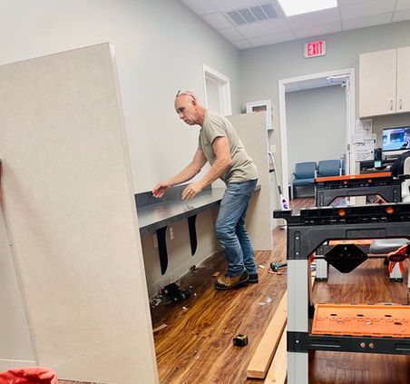 Man installs a counter in a room with wood floors. He's wearing jeans, a t-shirt, and work boots. Sawhorses are nearby.
