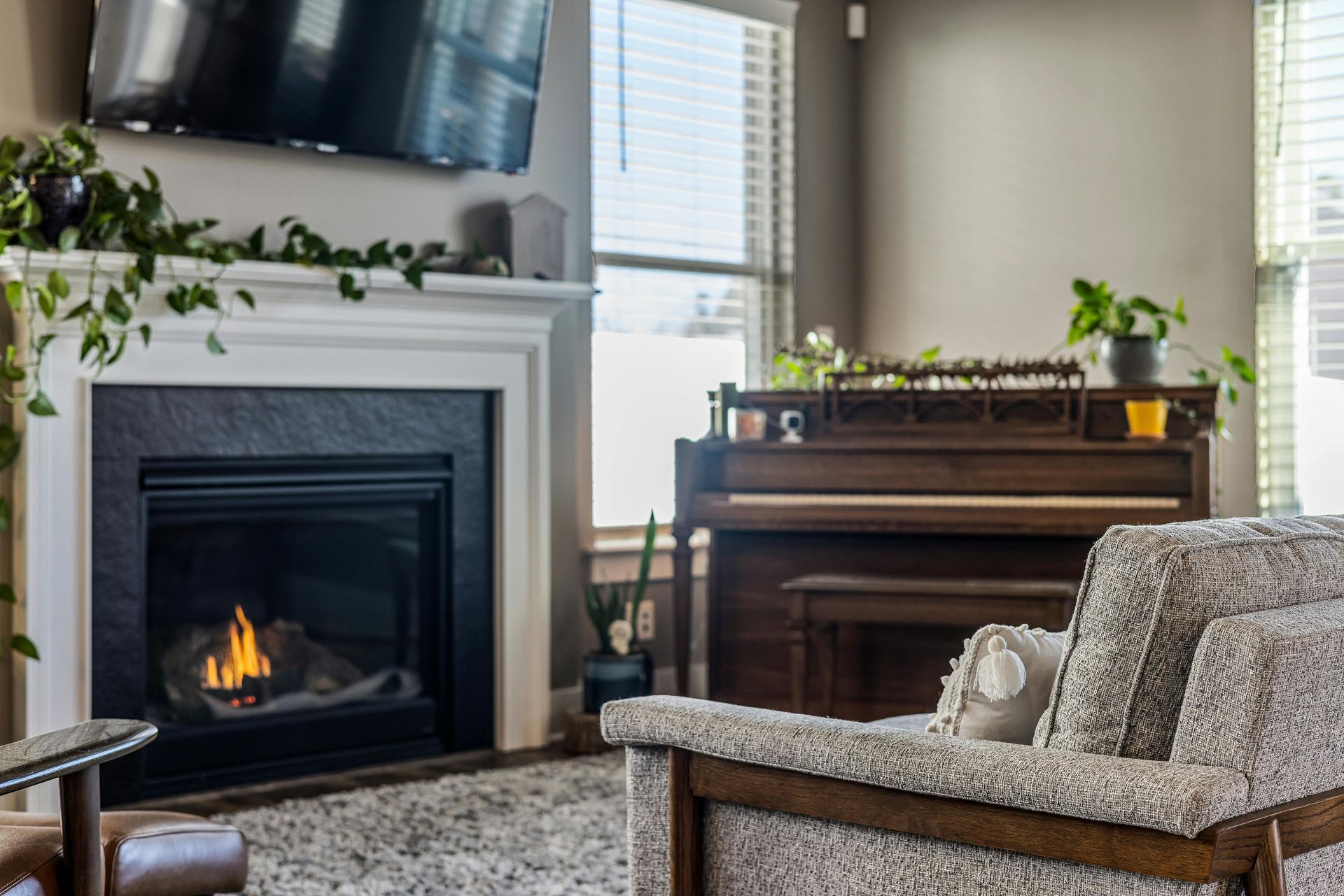 Cozy living room with fireplace, piano, and armchair; plants on mantel and piano top; window in background.