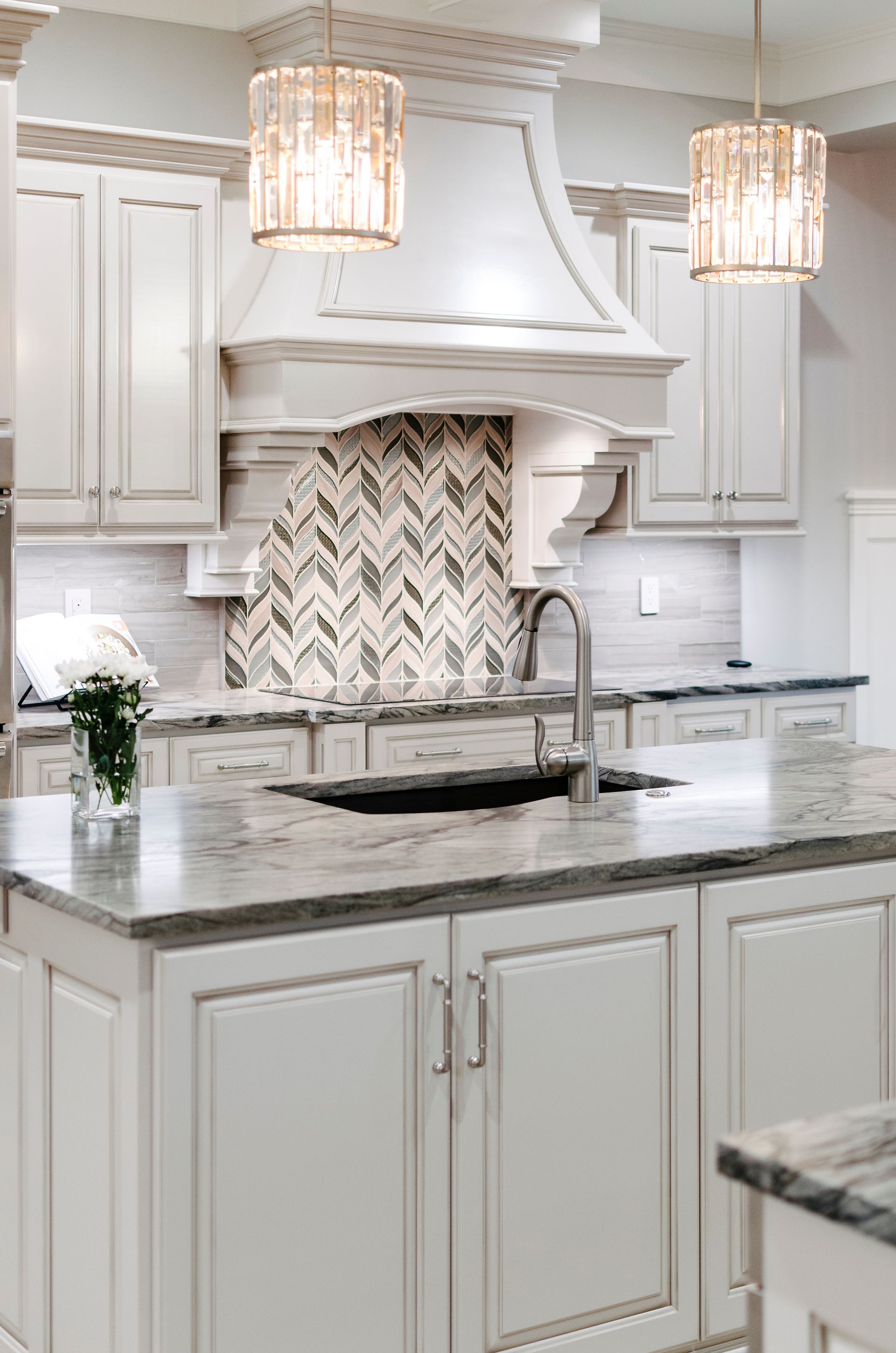 Elegant white kitchen with island, range hood, patterned backsplash, and crystal pendant lights.