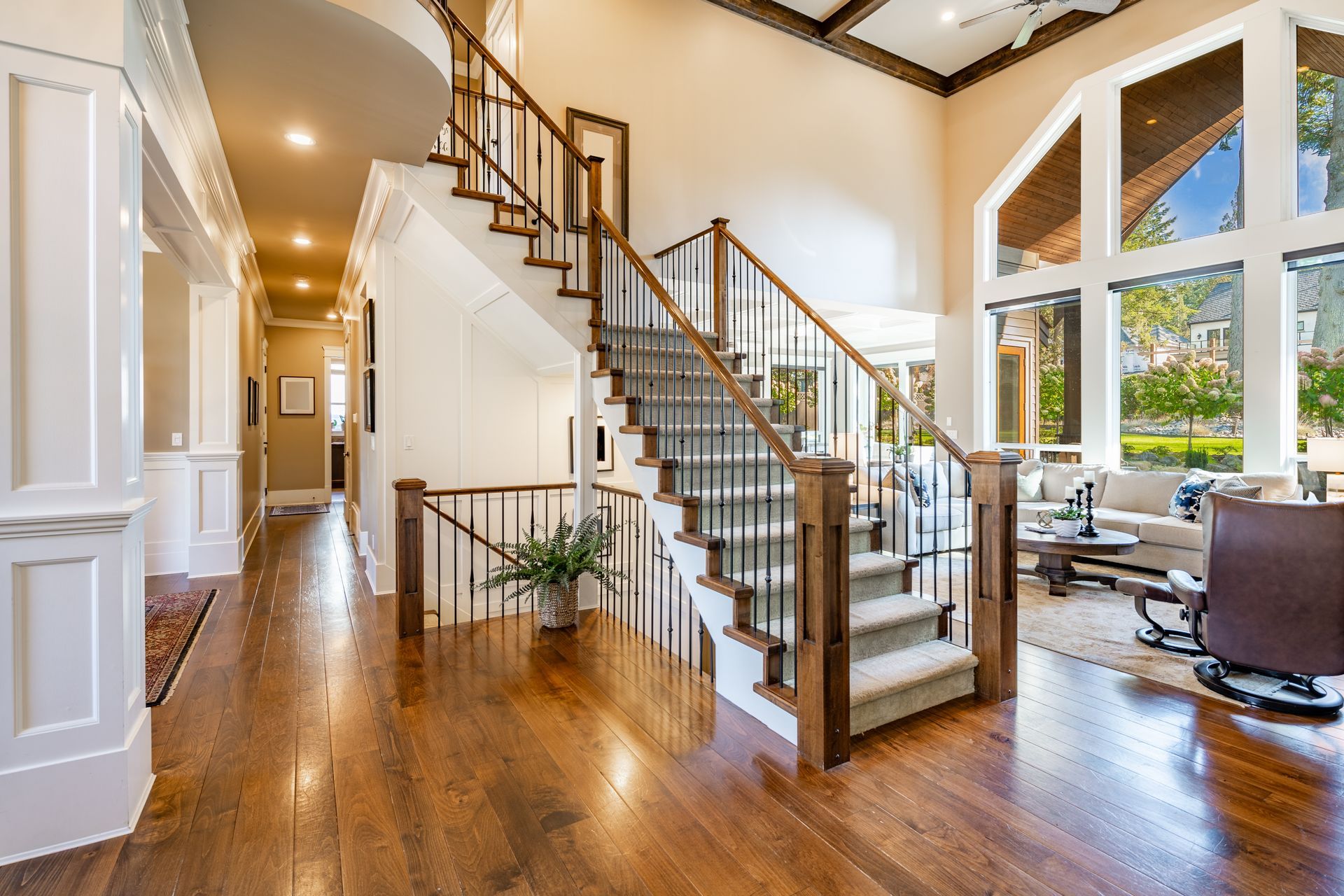 Wooden staircase in a bright foyer with hardwood floors, leading to a living room with tall windows.