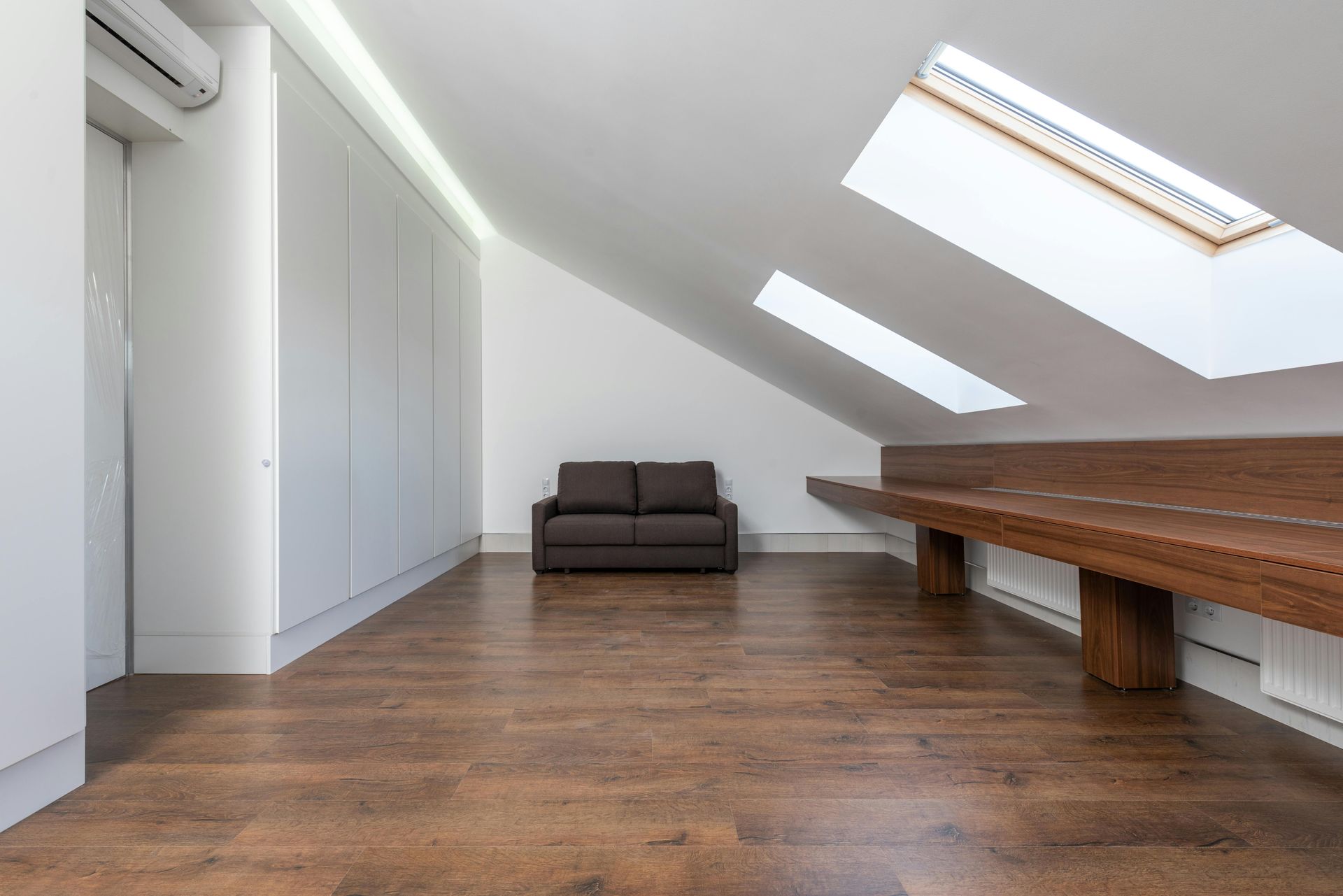 Attic room with brown sofa, wood floor, built-in white cabinets, skylights, and a long wooden bench.