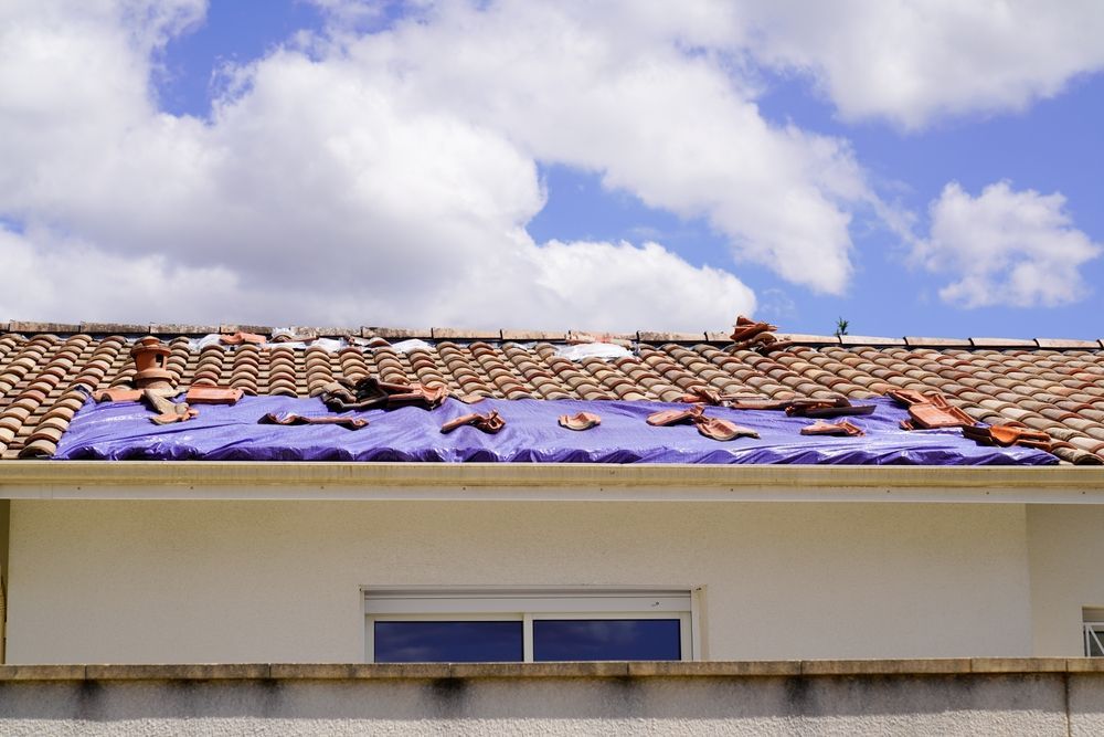 Damaged tile roof covered with a purple tarp. Blue sky with clouds in the background.