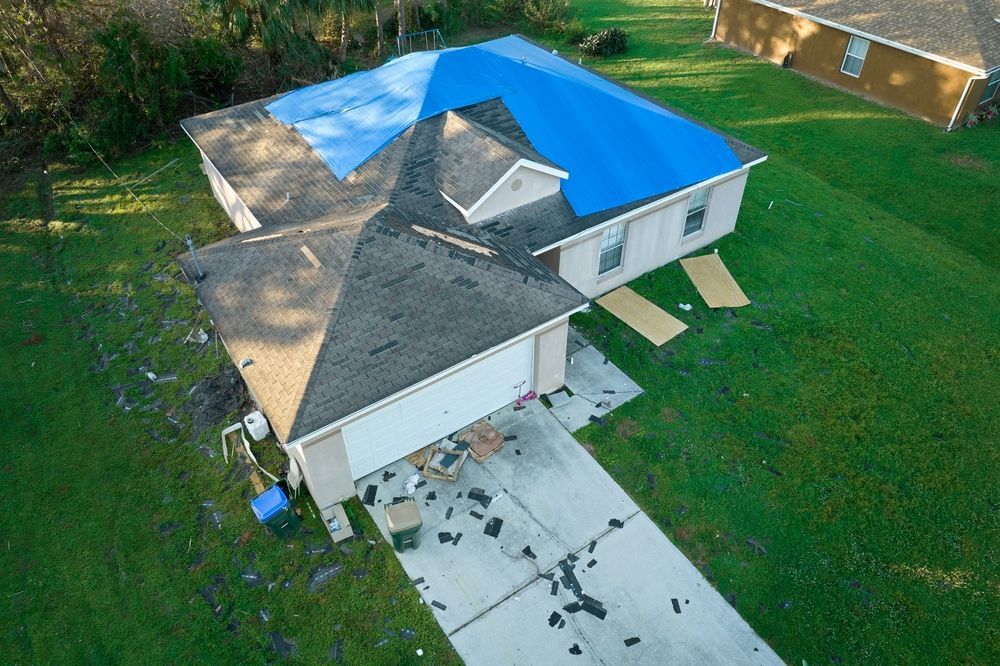 A house with a blue tarp on the roof after a storm, debris scattered on the driveway and lawn.