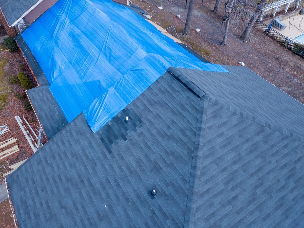 Blue tarp covers a damaged roof on a house. Dark gray shingles are visible in the foreground.