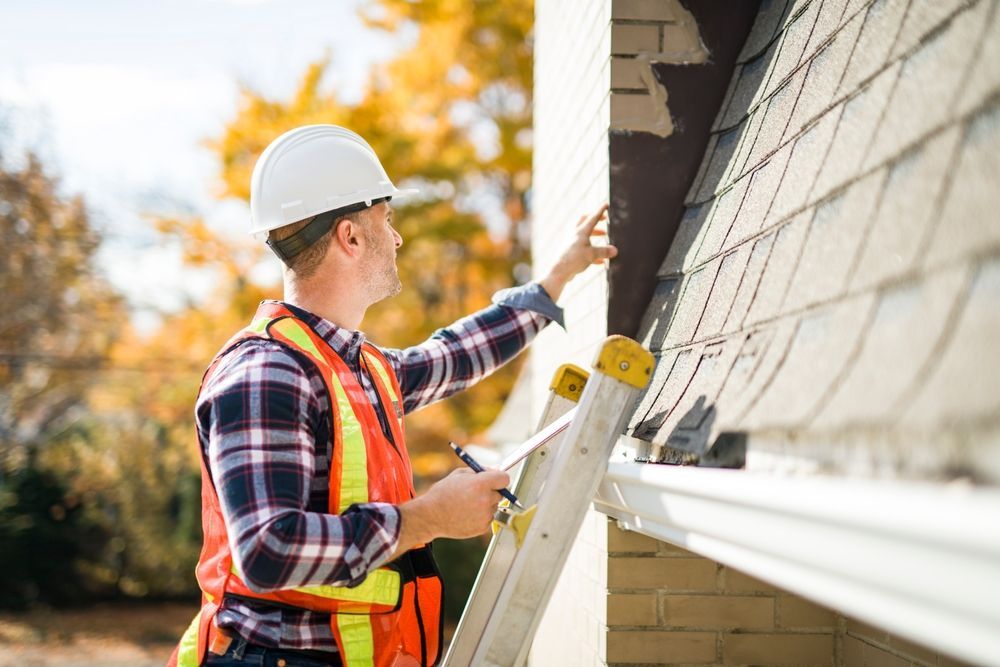Man in hard hat and vest inspecting a roof, standing on a ladder next to a house.