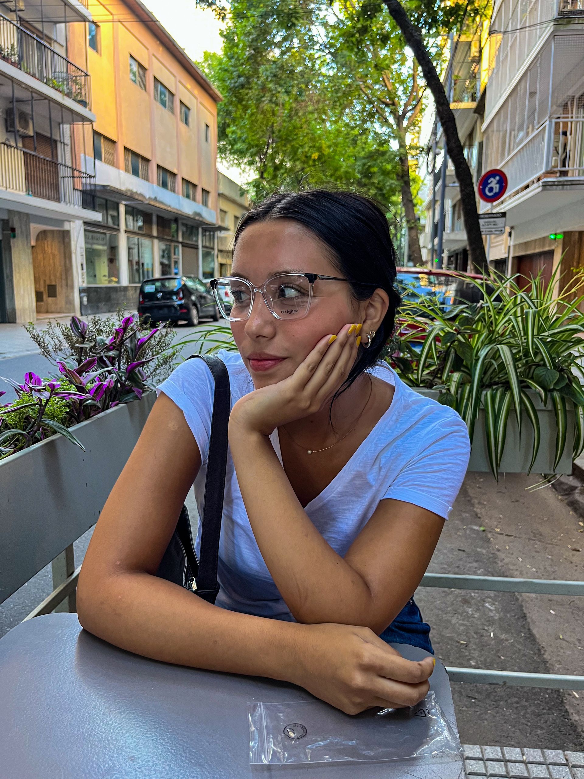 Mujer con gafas, apoyando la cara en la mano, sentada en una mesa al aire libre. Edificios al fondo.