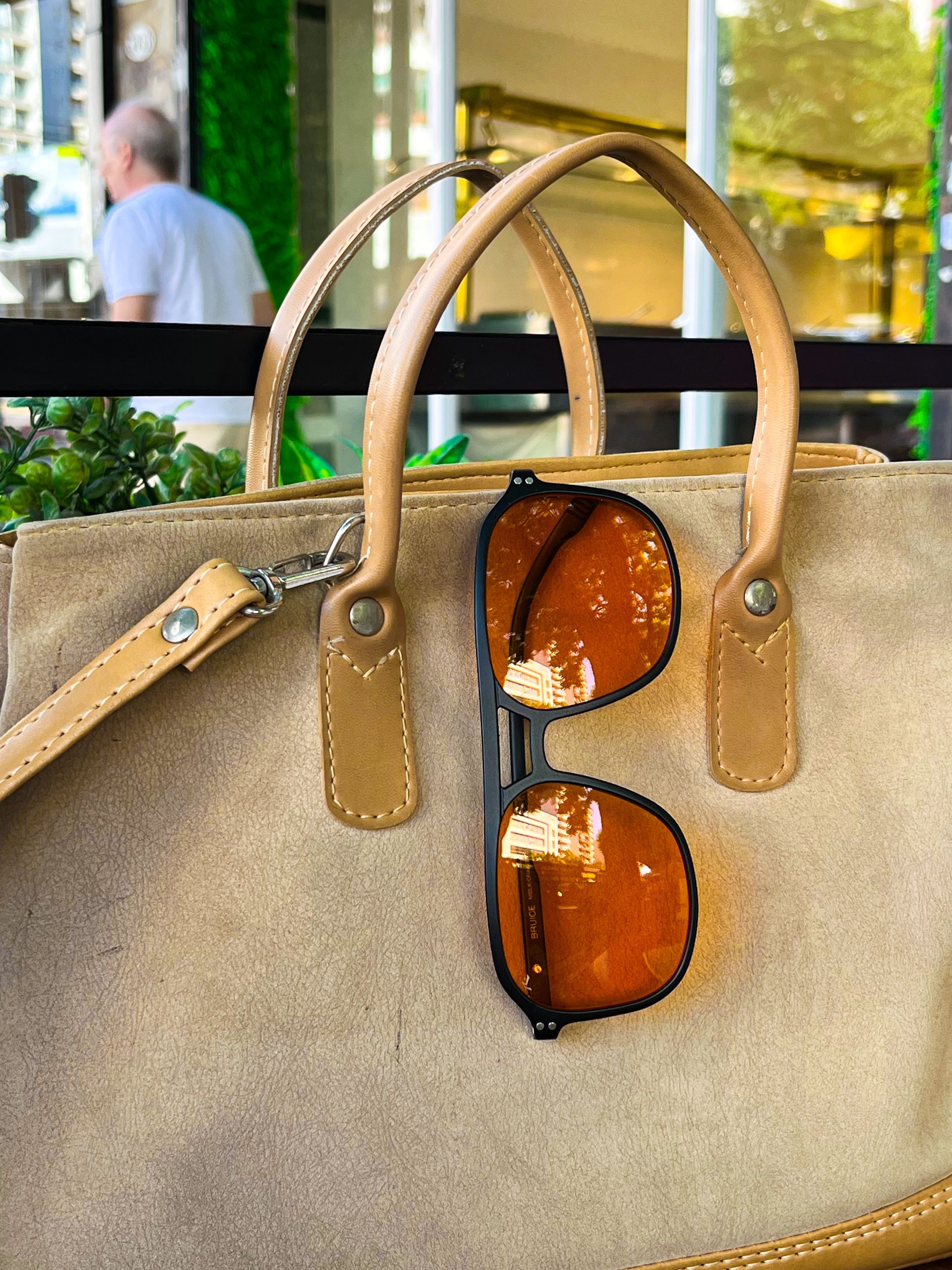 Bolso de mano de lona beige con detalles de cuero marrón y gafas de sol, apoyado sobre una mesa de madera.