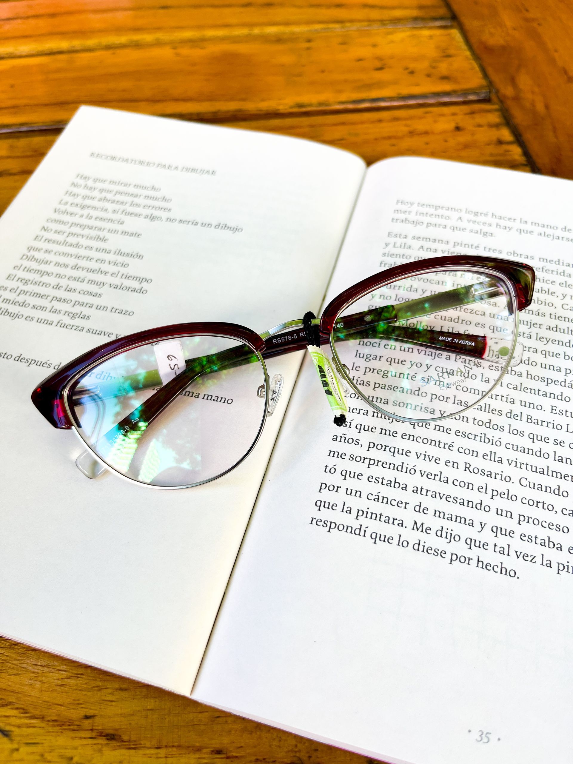 Unas gafas con montura burdeos y plata descansan sobre un libro abierto, sobre una mesa de madera.