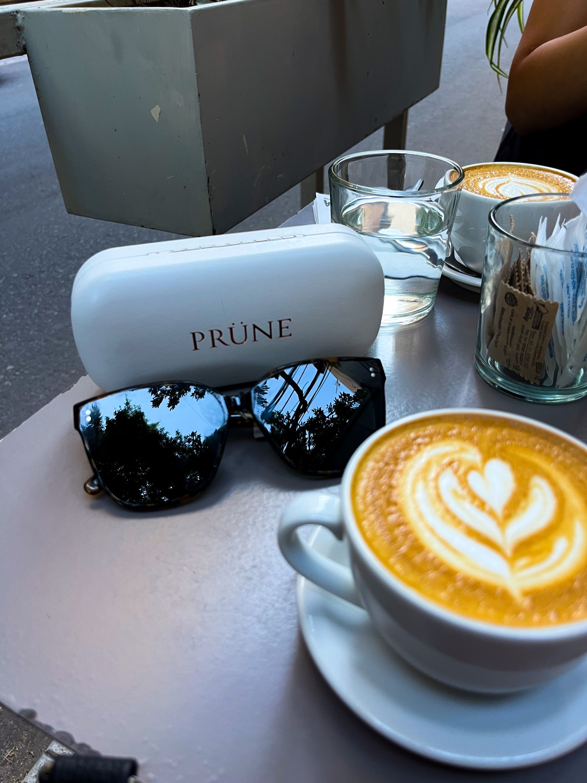 Gafas de sol, una taza de café con arte latte y otros artículos de cafetería sobre una mesa.