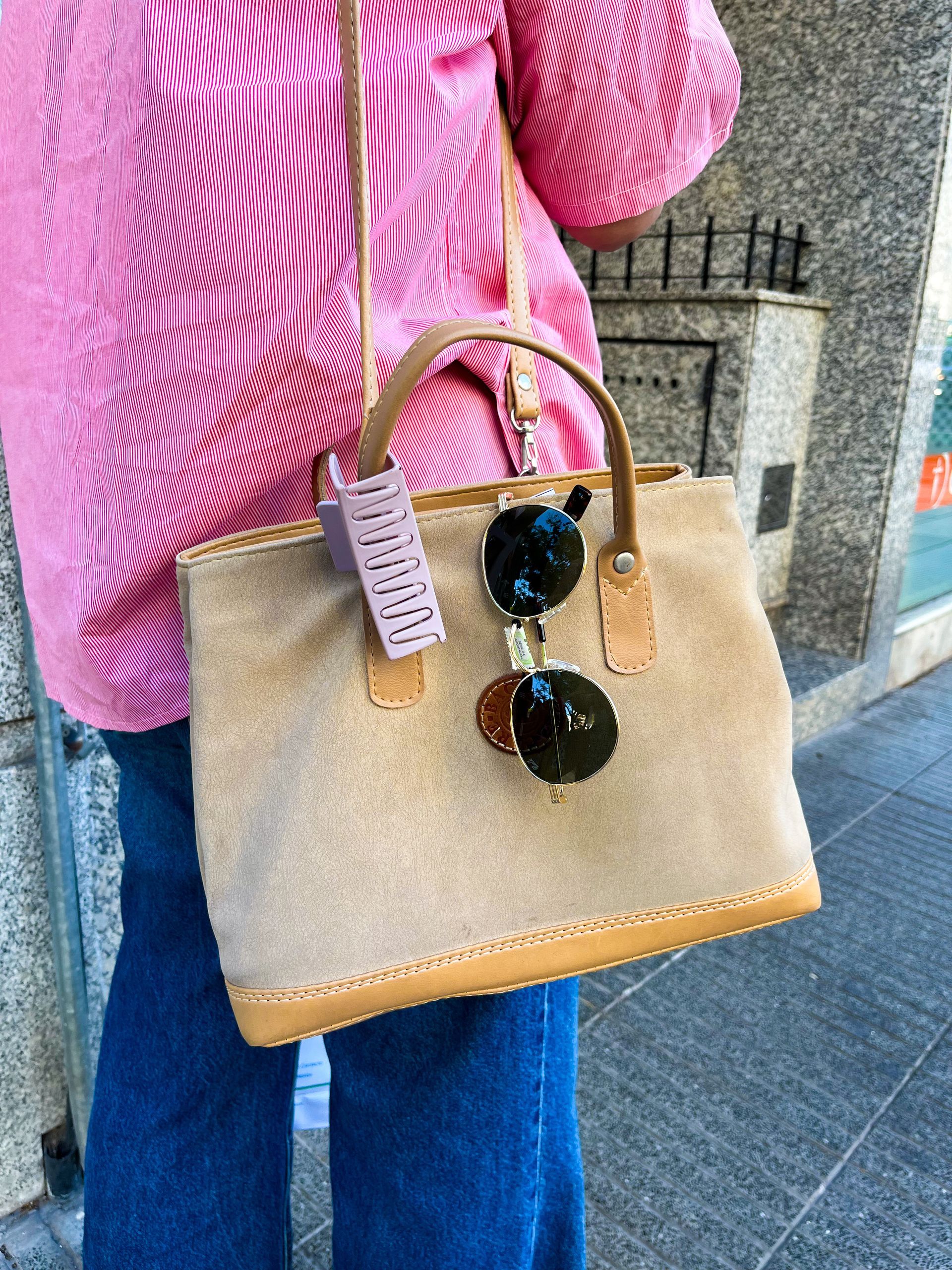 Persona que viste una camisa rosa y pantalones vaqueros azules sosteniendo un bolso de gamuza color tostado con gafas de sol