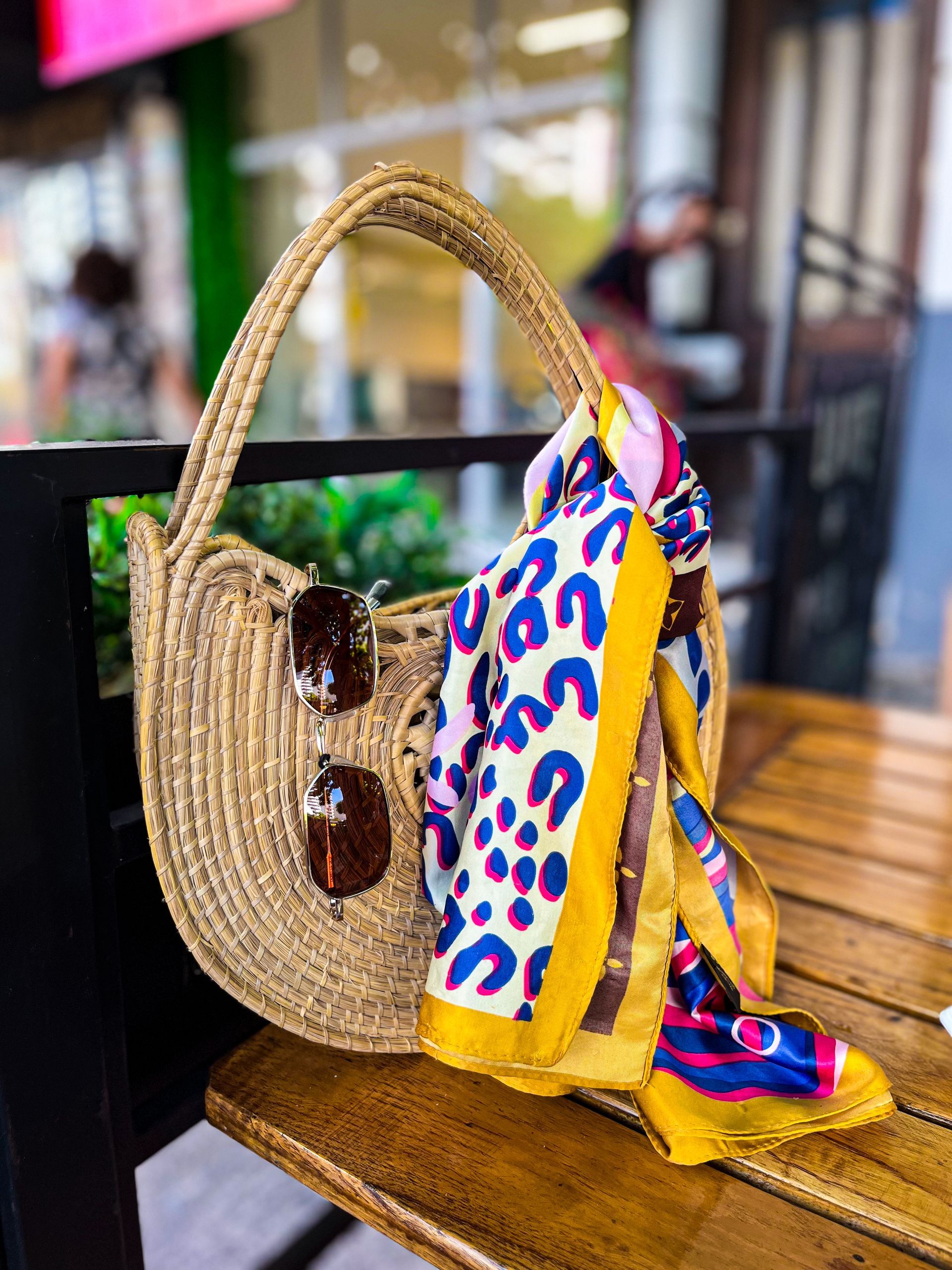 Gafas de sol colgadas en un bolso tejido junto a una bufanda colorida; vegetación en el fondo.