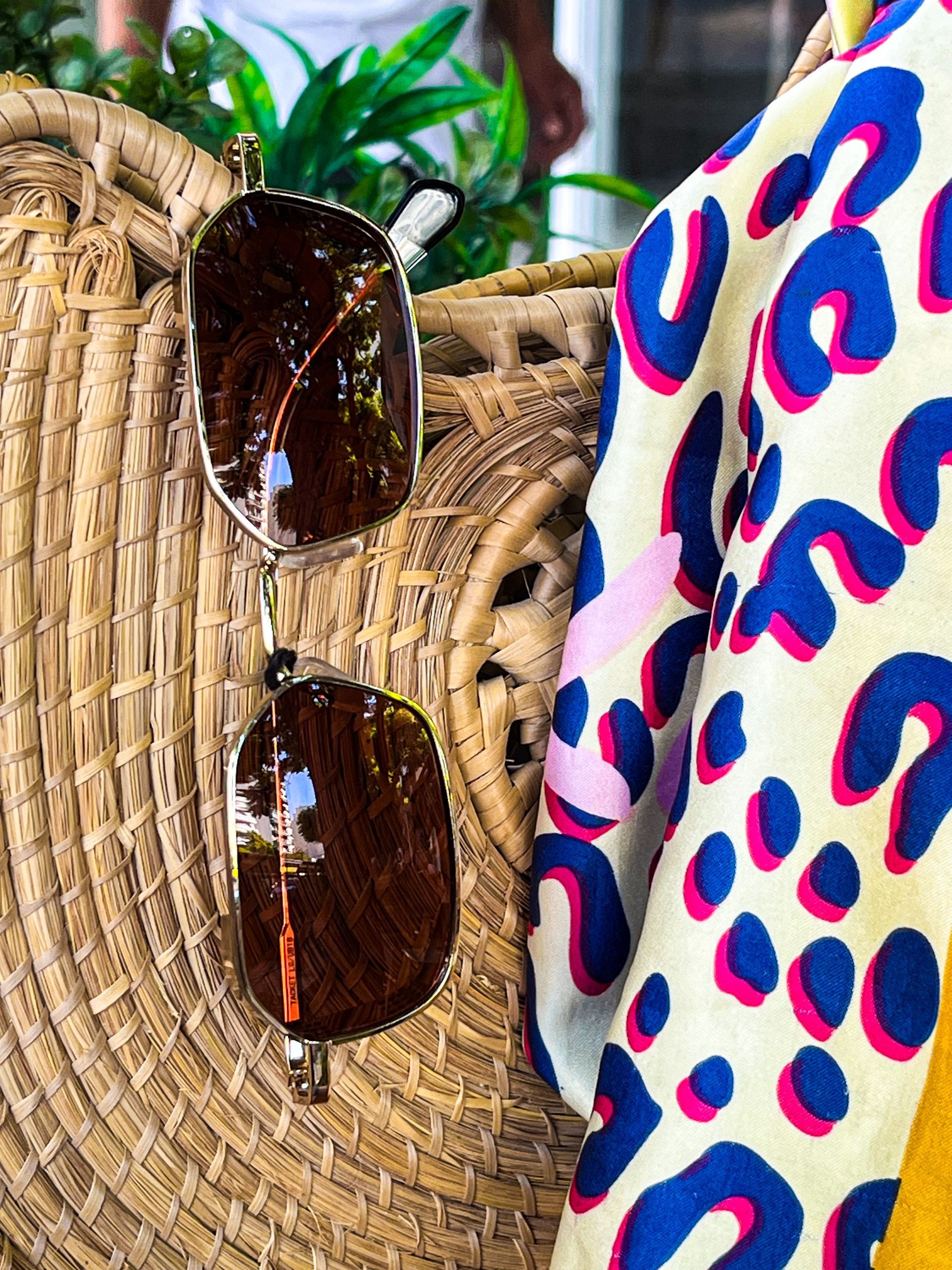 Gafas de sol colgadas en un bolso tejido junto a una bufanda colorida; vegetación en el fondo.