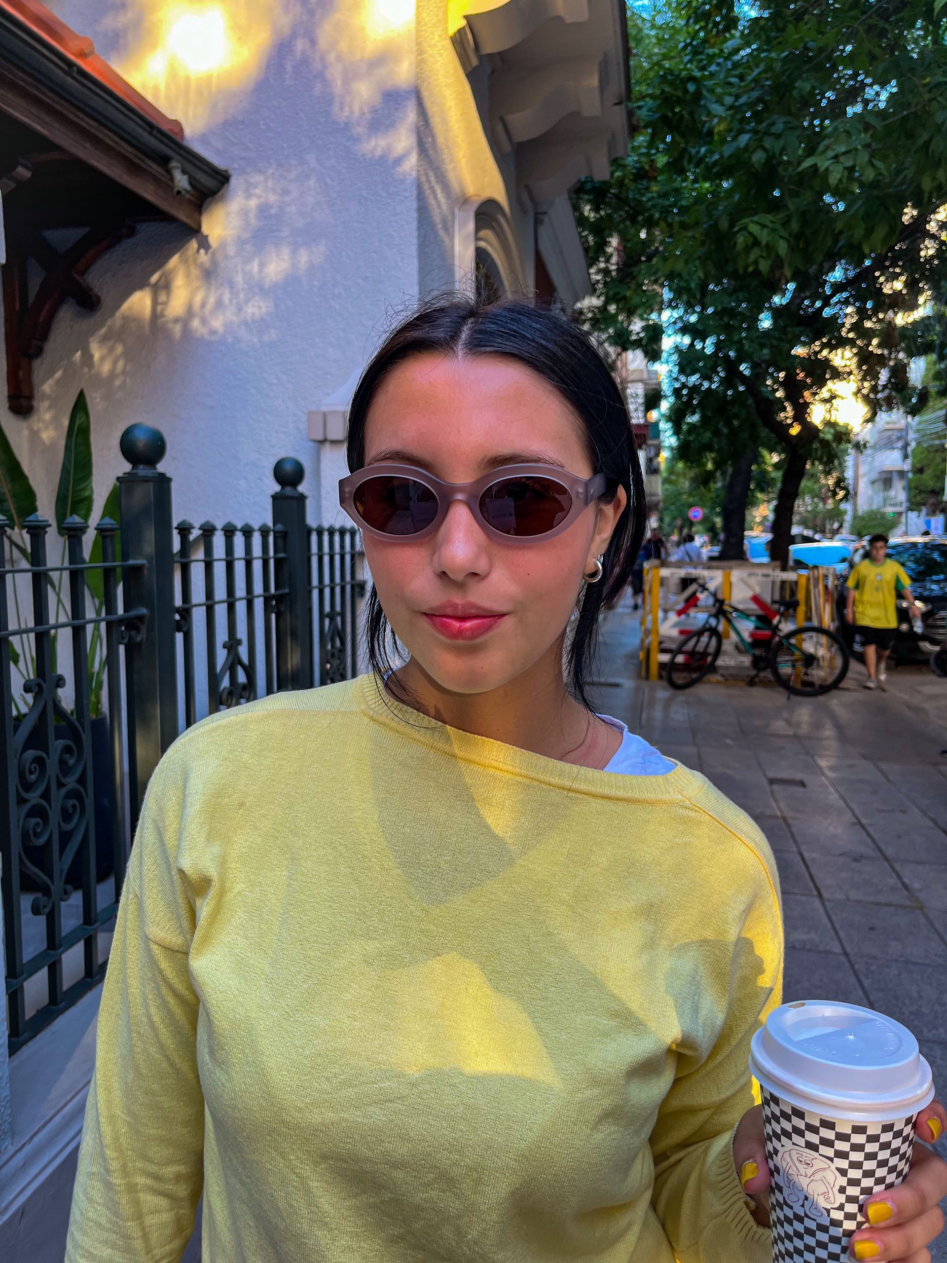 Mujer con gafas de sol y taza de café al aire libre, vistiendo un suéter amarillo.