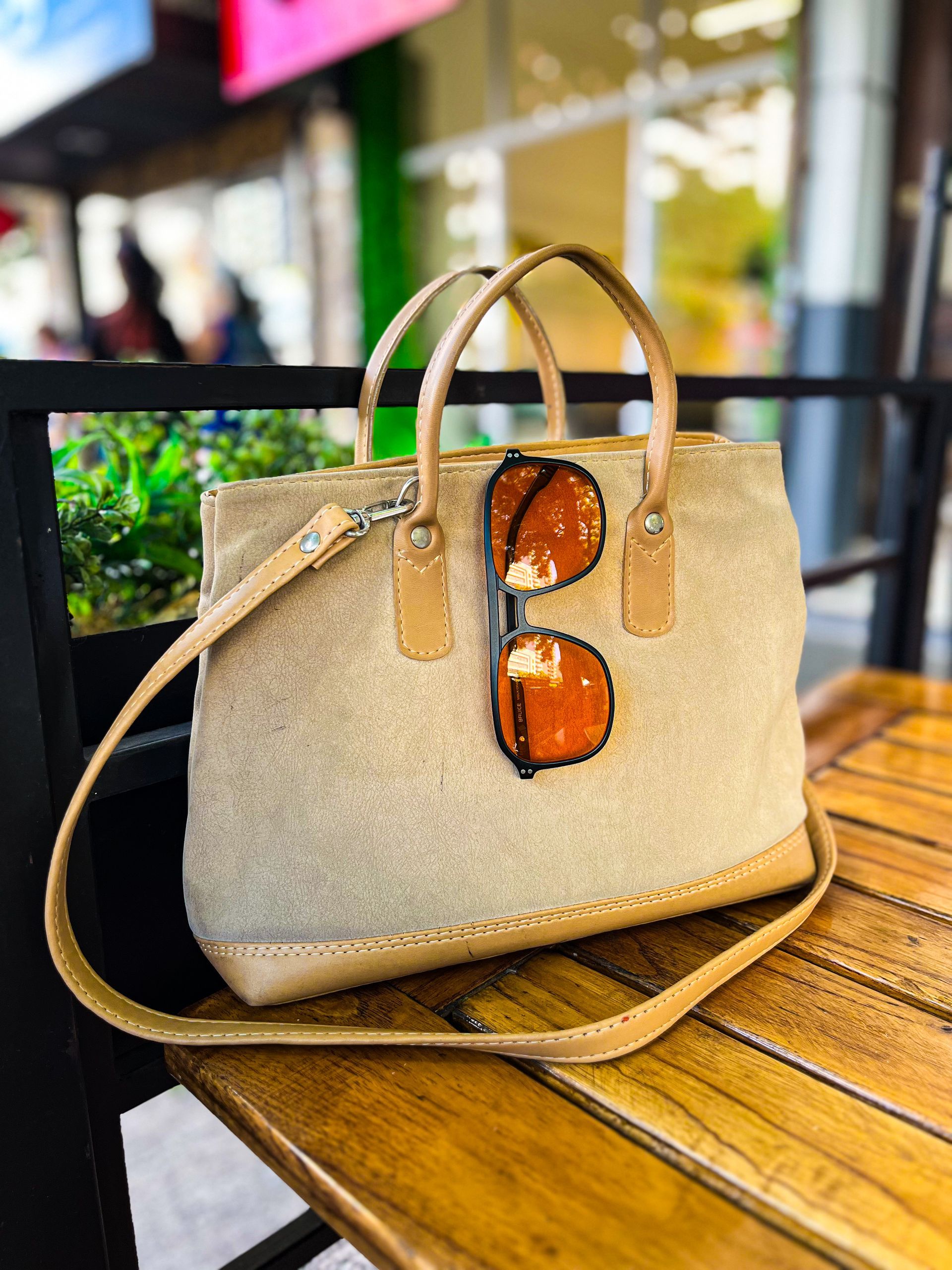 Bolso de mano de lona beige con detalles de cuero marrón y gafas de sol, apoyado sobre una mesa de madera.