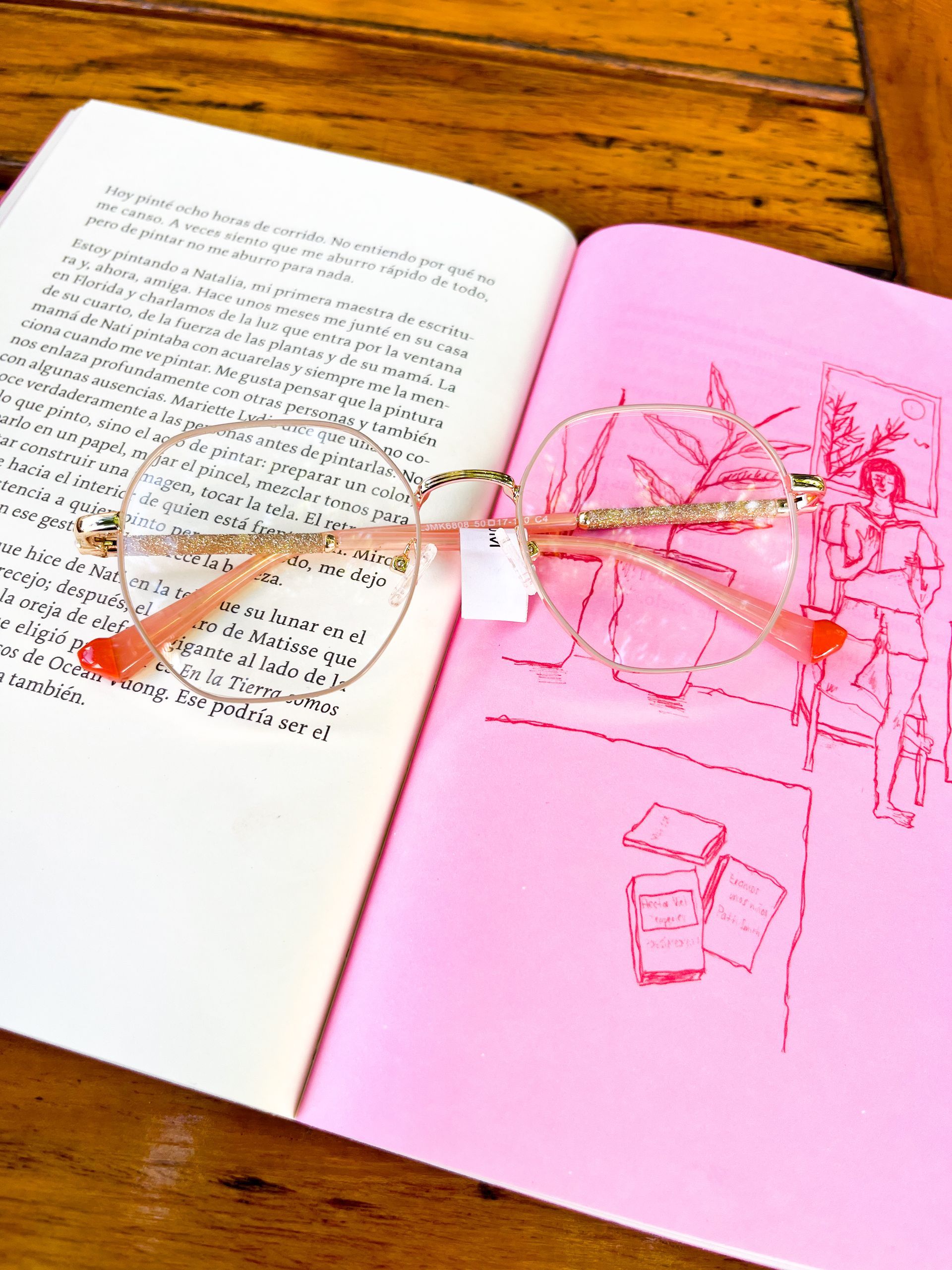 Gafas sobre un libro abierto, un lado con texto impreso y el otro rosa con boceto.