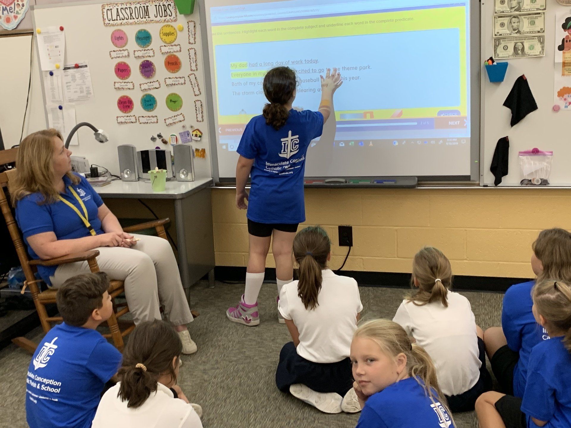 A girl wearing a blue shirt that says 'IC' on it is giving a presentation to a group of children