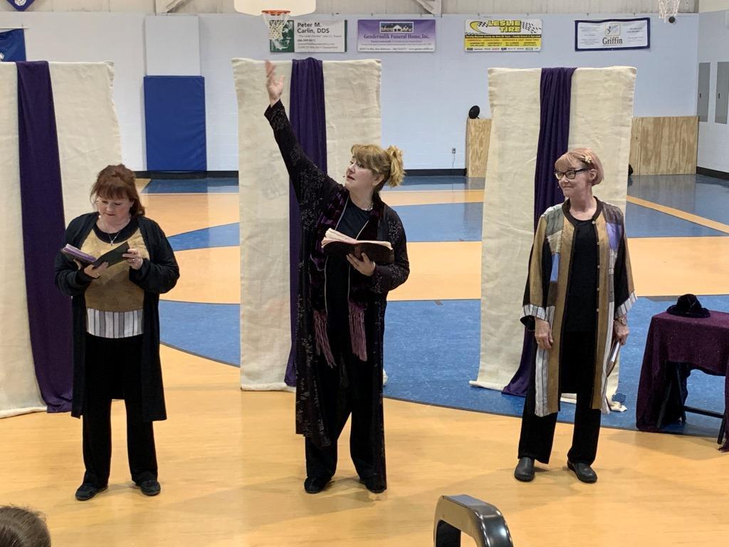 Three women are standing in a gym and one is holding a book
