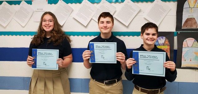 Three students in school uniforms are holding certificates in front of a wall.