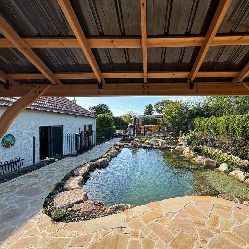 A Pond Is Surrounded By Rocks Under A Wooden Roof — Premier Bricks, Blocks & Pavers In Heatherbrae, NSW