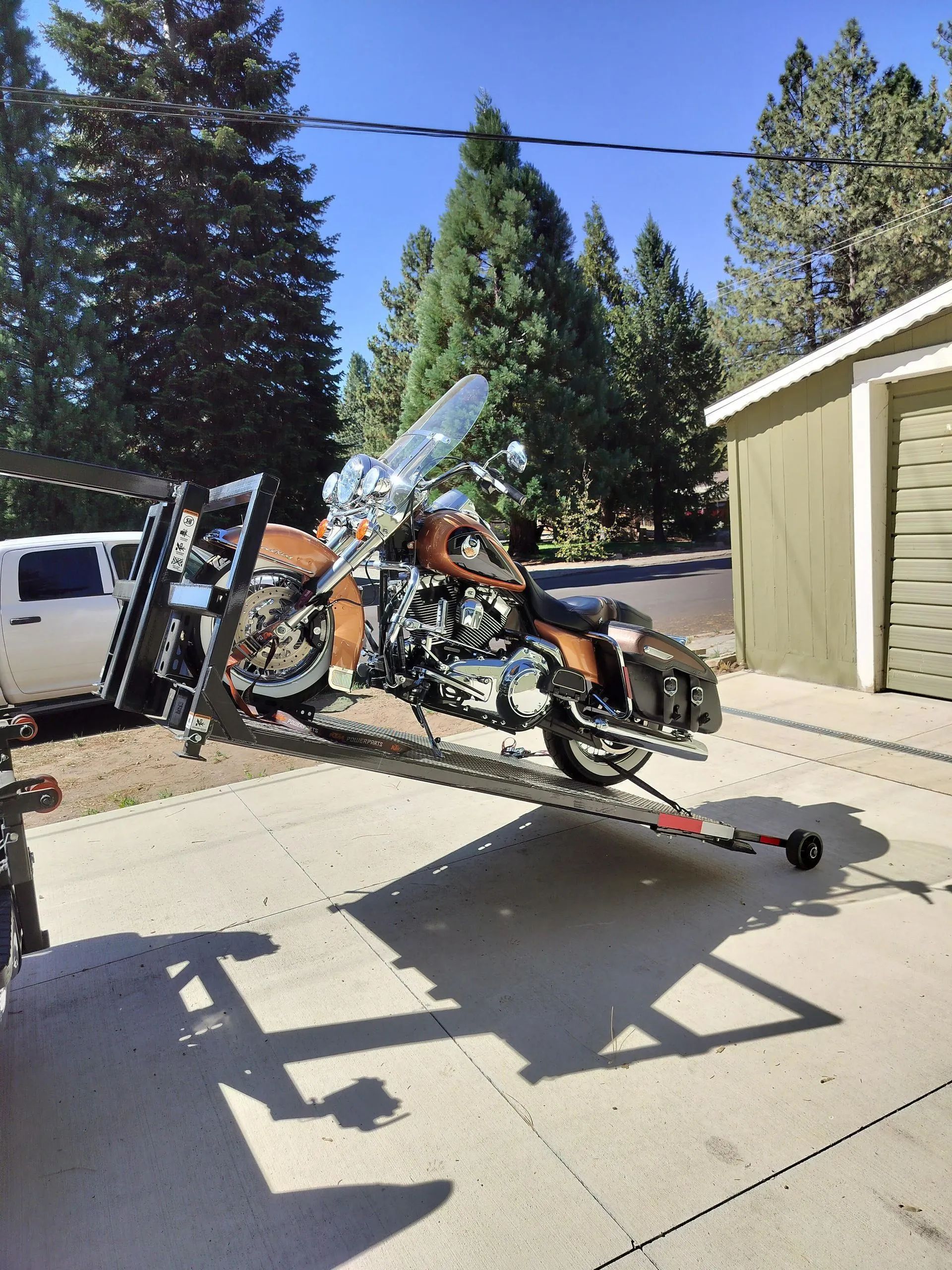 Motorcycle loaded onto a trailer in a driveway with trees and a garage in the background on a sunny day.