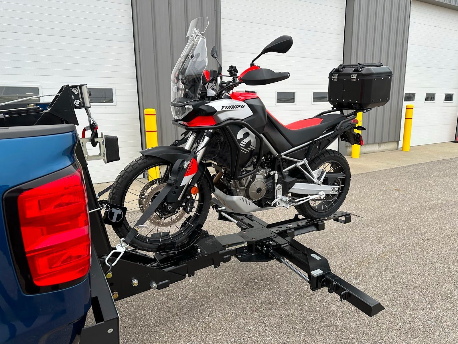 Motorcycle mounted on a hitch carrier attached to the back of a blue truck, outside a building.