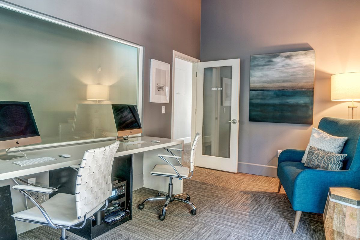Interior community workspace with two iMacs on a long white desk, white chairs, a frosted glass divider, and a blue sofa. Explore 1 and 2 bedroom apartments and townhomes in Larkspur, CA with modern floor plans and a convenient location just a 30-minute drive from San Francisco.