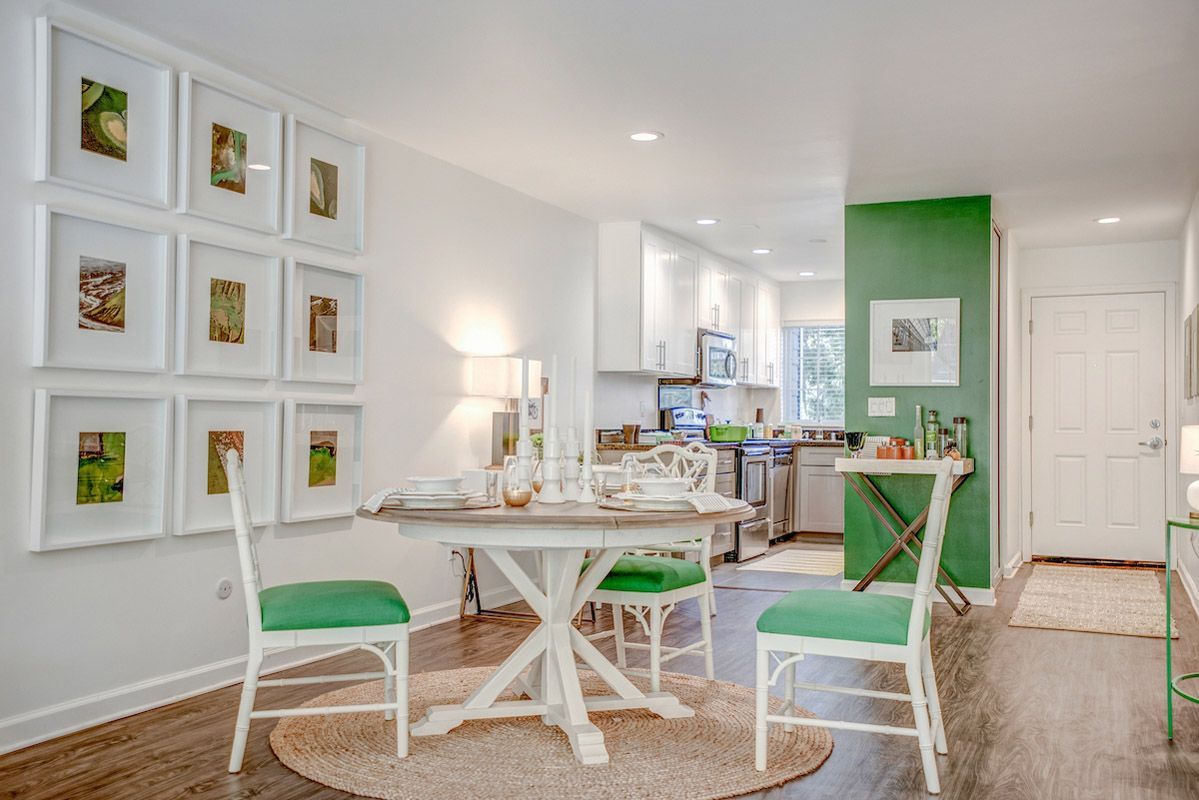 Open dining area in a modern apartment: round table, white chairs with green cushions, kitchen in background. View our 1- and 2-bedroom homes and townhomes at Finch at Larkspur in scenic Larkspur, CA.  