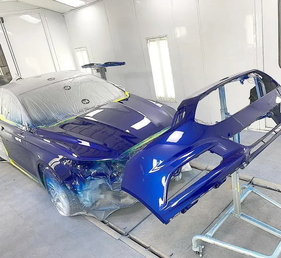 Blue car in a paint booth with its bumper removed, ready for painting.