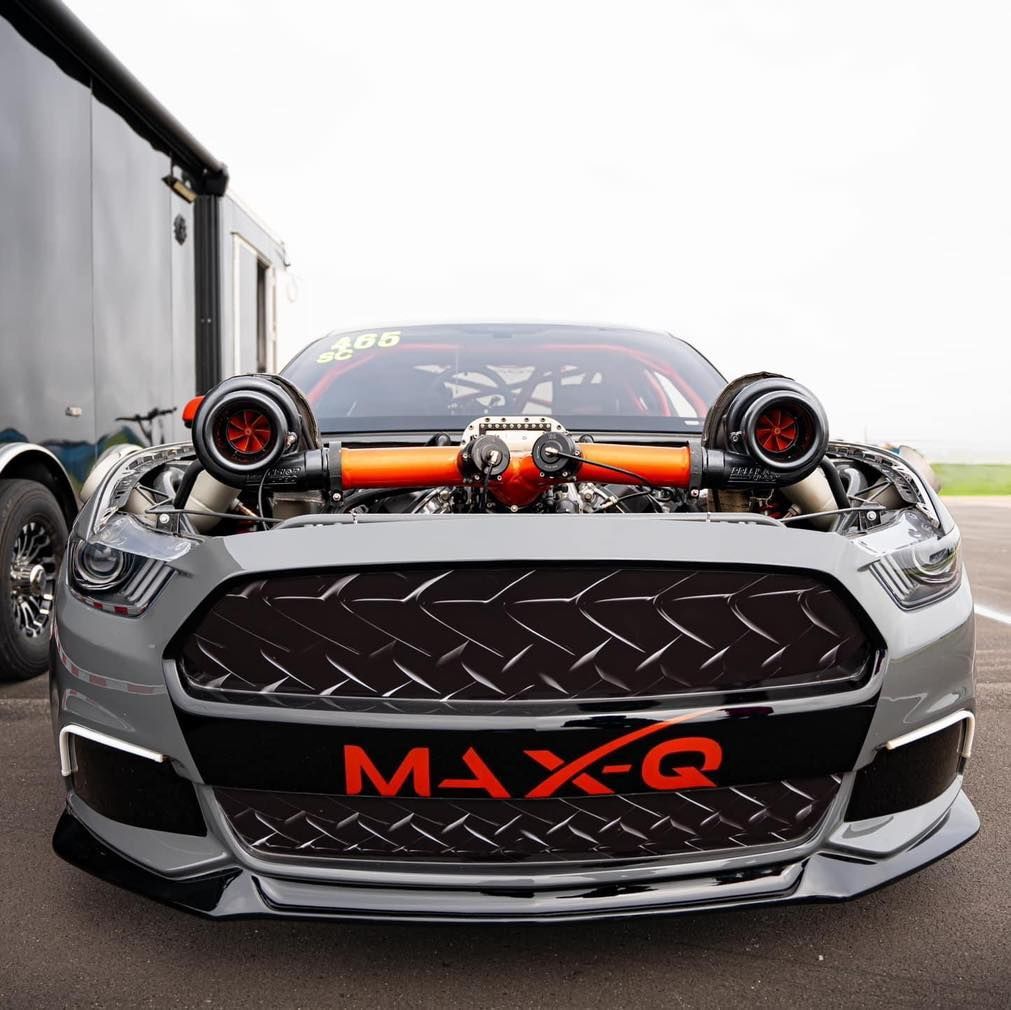 Gray race car with large turbos, orange piping, and "MAX-Q" logo in front of a trailer.