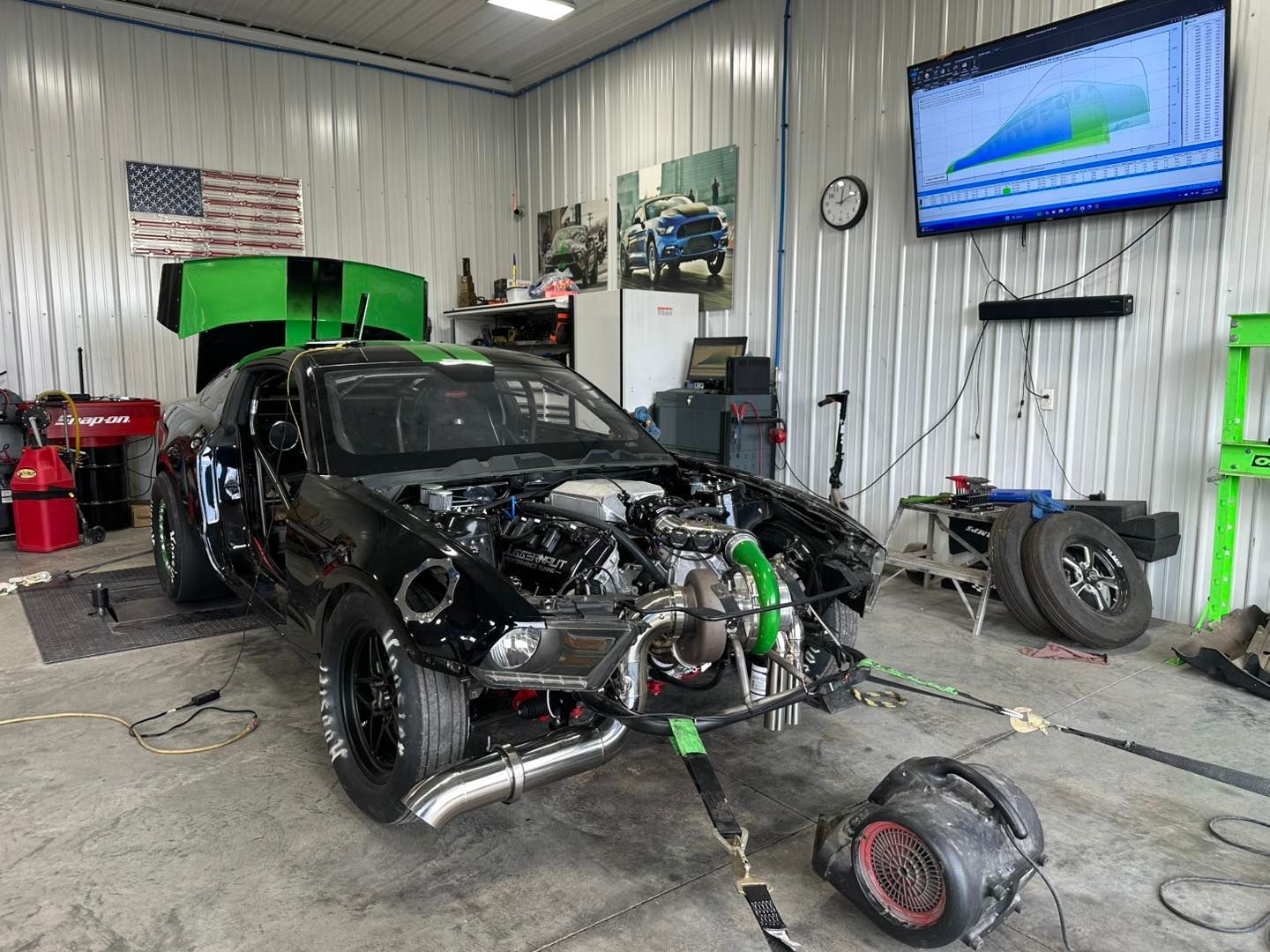 Black car on a dyno in a garage with a large turbocharger and connected to a monitor displaying engine performance.