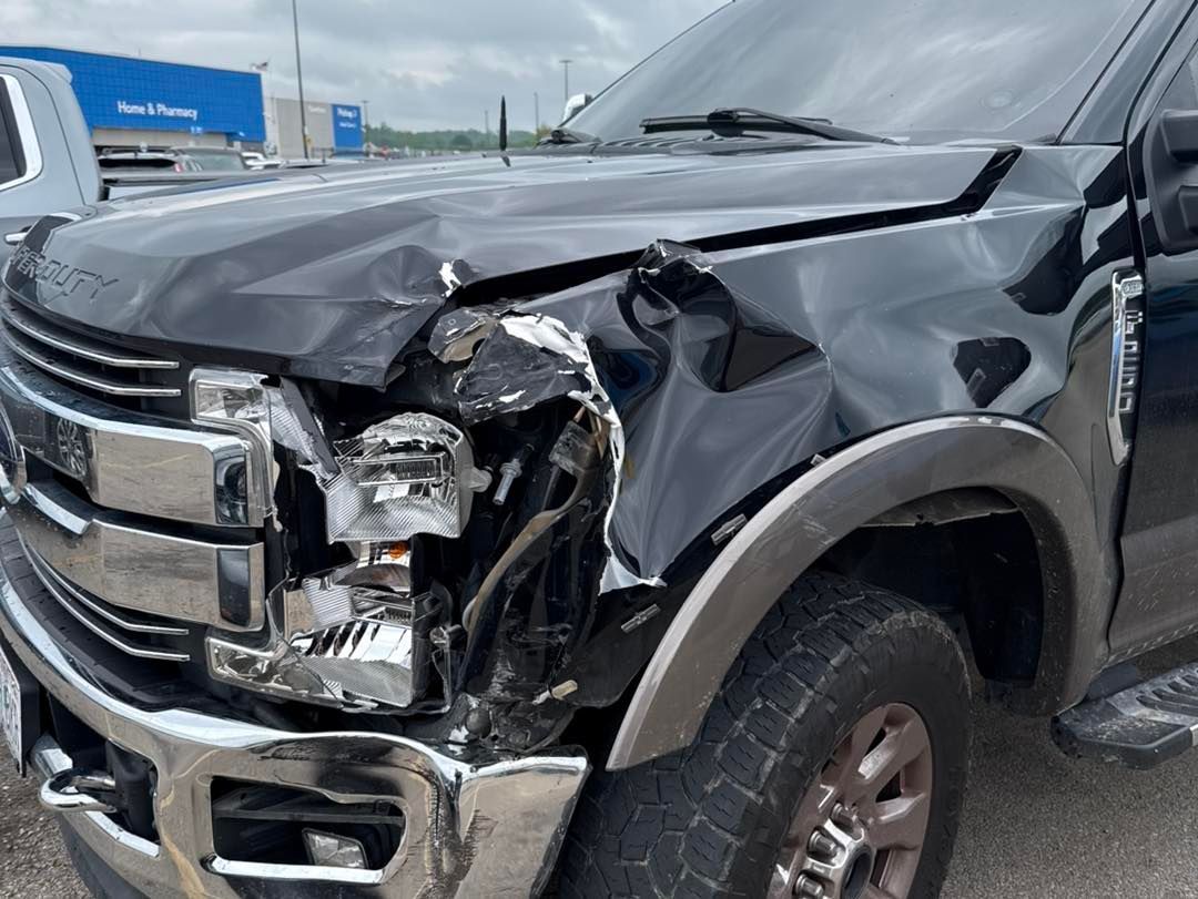 Damaged black Ford truck front end with crushed headlight, crumpled hood and fender.