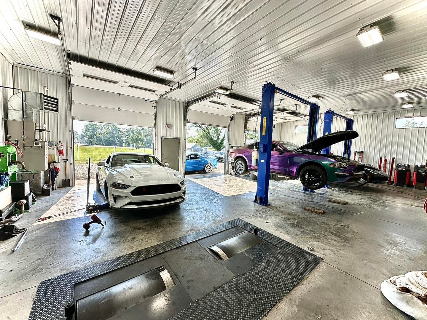 Cars in a garage. A white Mustang sits on a lift. Another car is lifted with its hood open. Garage doors are open.
