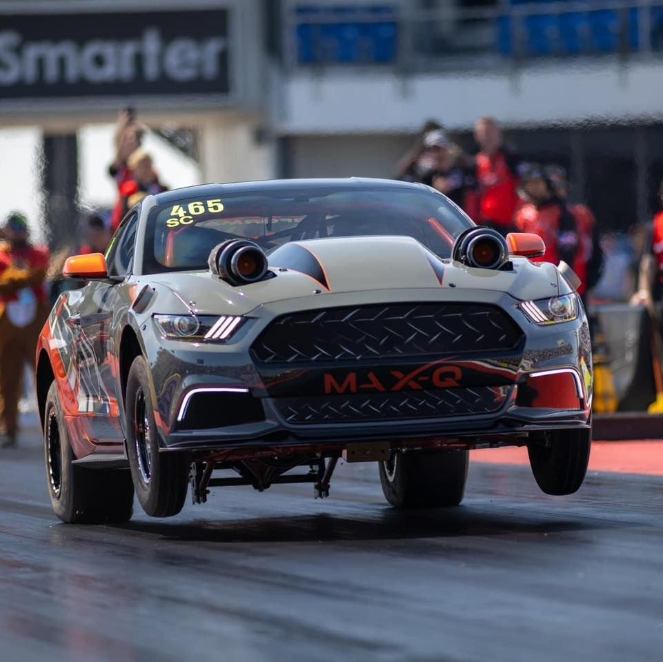 A gray and orange drag racing Mustang with front wheels off the ground, racing at a track.