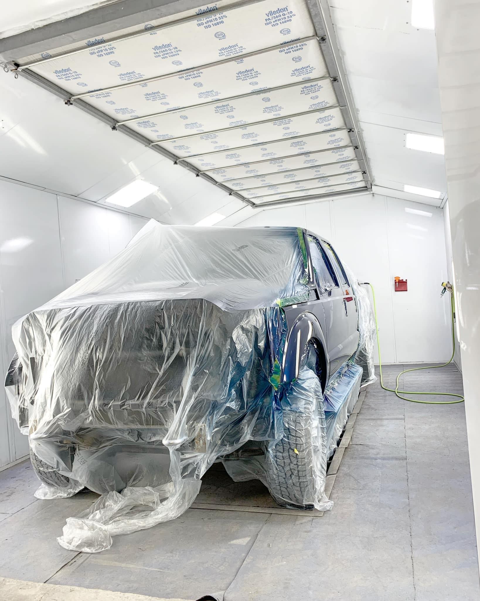 SUV covered in plastic wrap inside a spray paint booth with overhead lights.