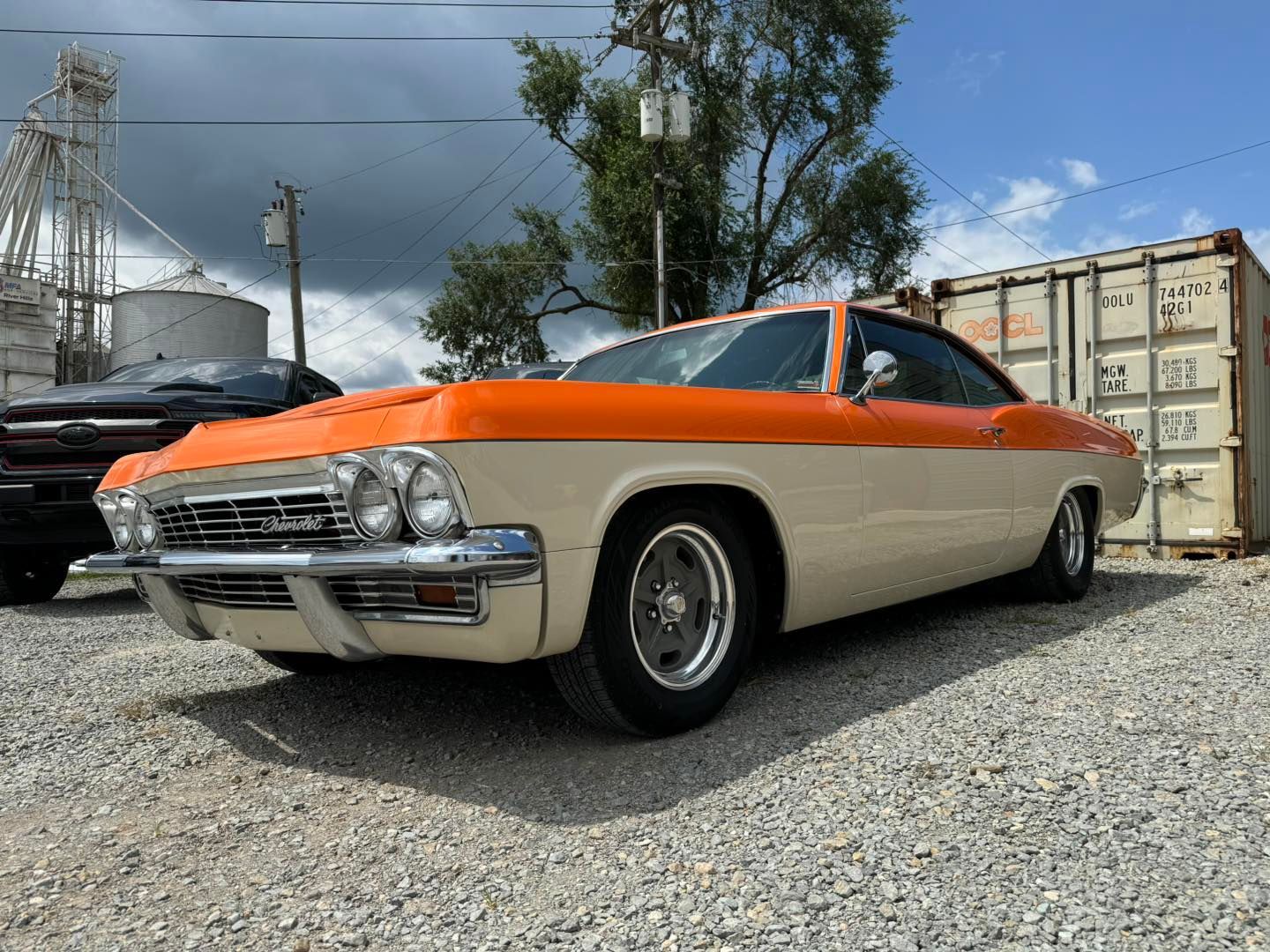 Two-tone orange and cream classic car parked on gravel, with shipping containers and sky in background.