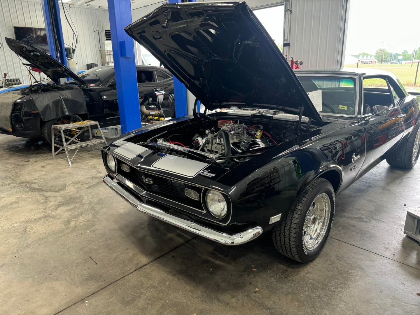 Black classic car with hood open in a garage, another car in the background.