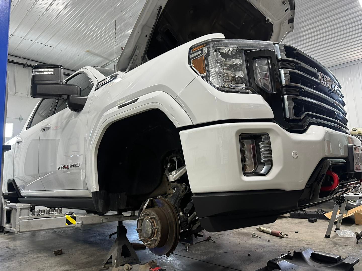 White GMC truck on a lift with hood open, wheel removed, in a repair shop.