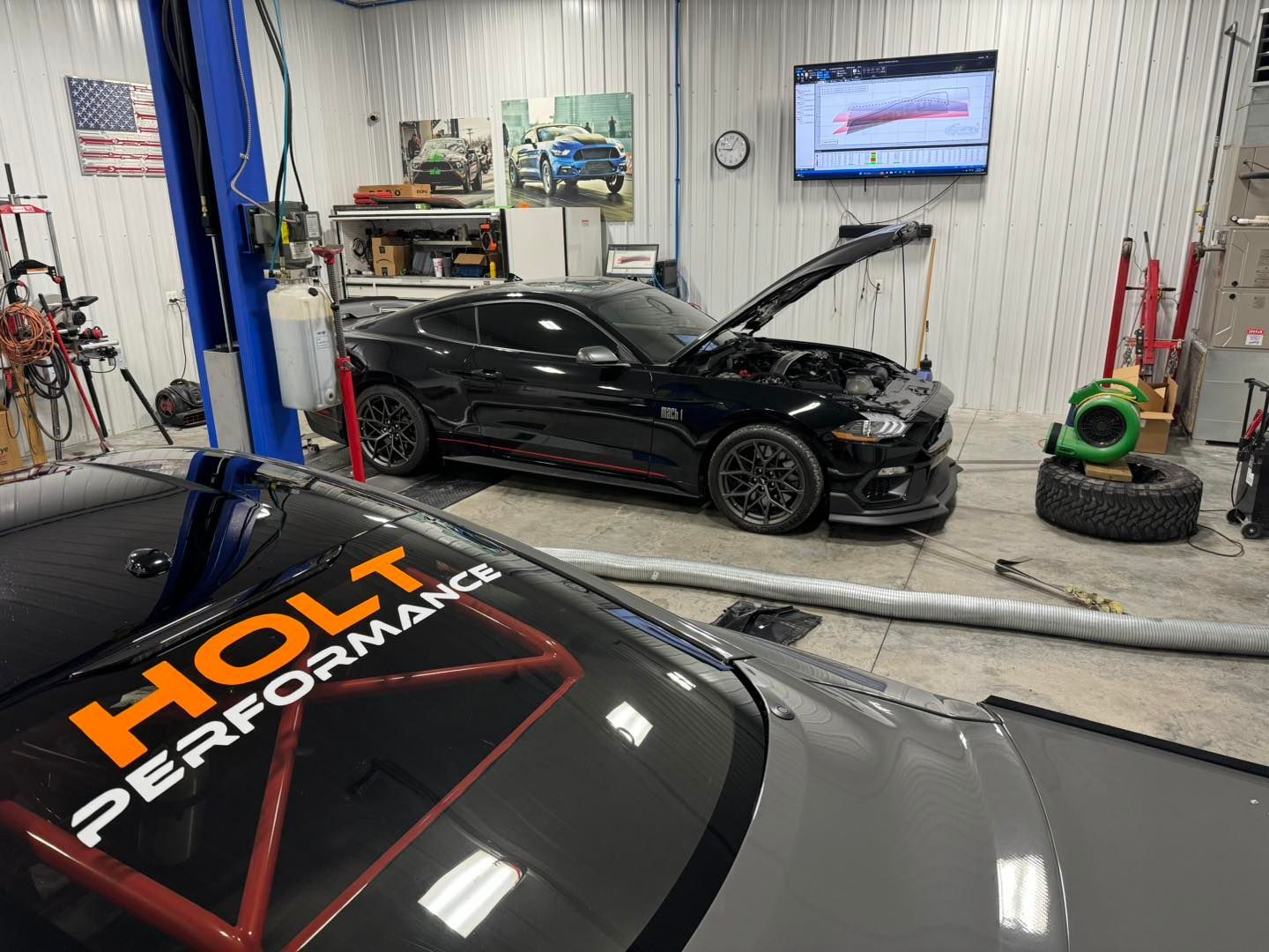 A black Ford Mustang with the hood open in a garage, another car in the foreground with "HOLT PERFORMANCE" on the windshield.