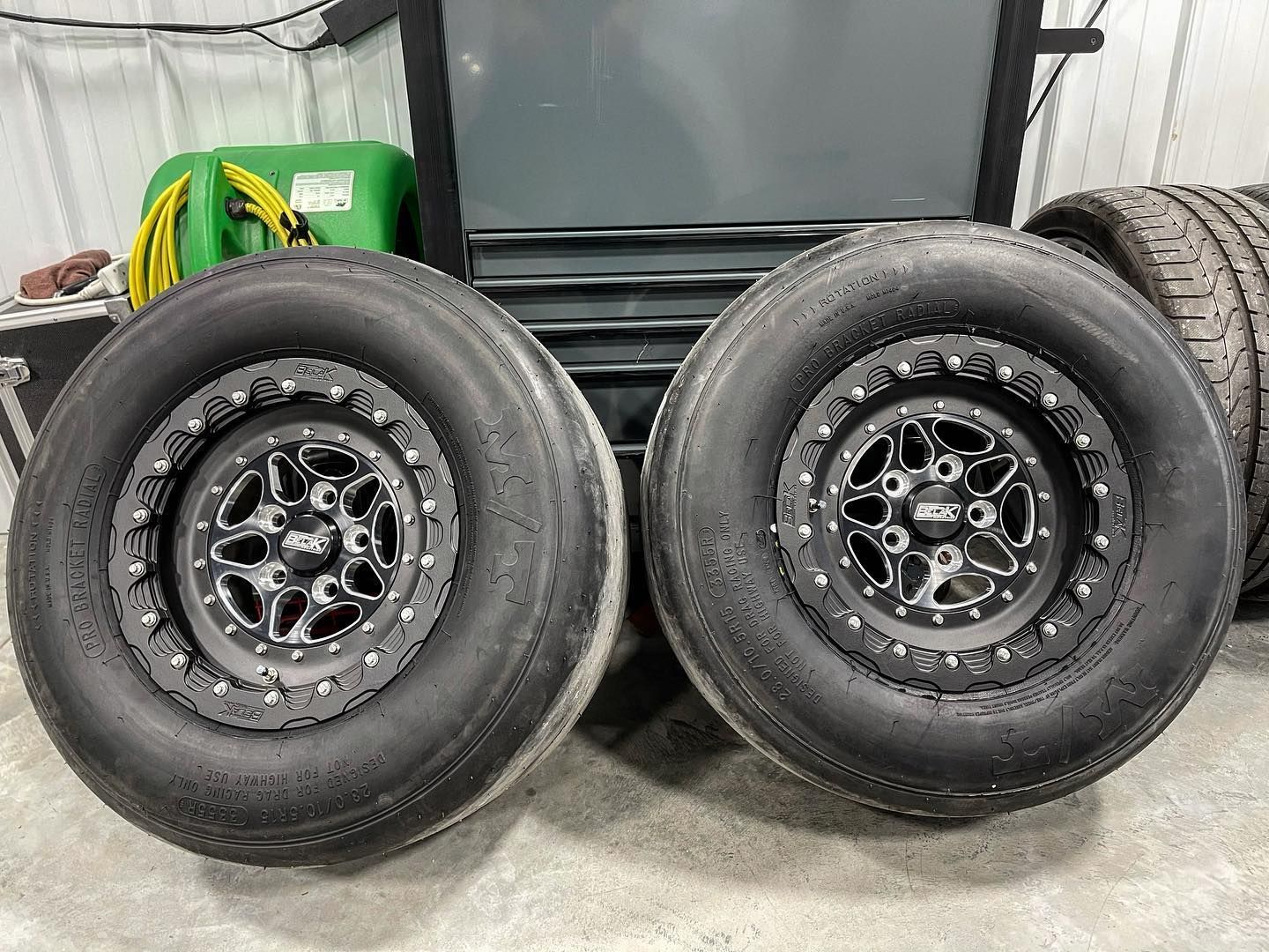 Two black drag racing tires with black rims on a garage floor.