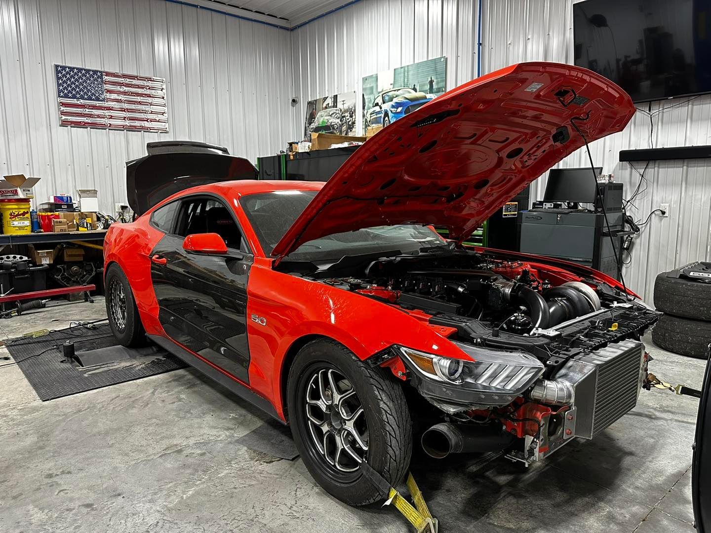 Red and black Mustang with the hood open in a garage, being worked on.