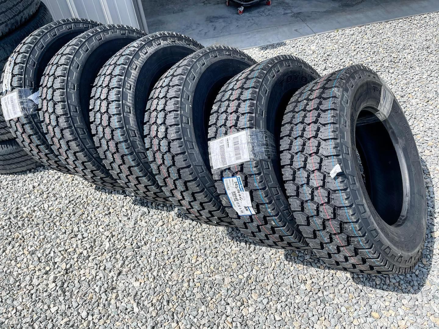 Seven new black truck tires stacked on gravel.