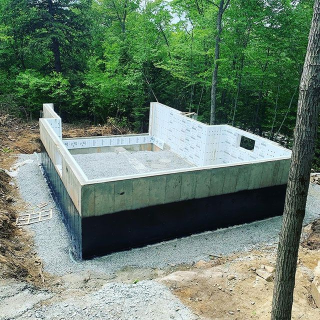 Fondations d'un bâtiment en construction, entourées de gravier, dans une zone boisée. Les murs extérieurs sont en béton.