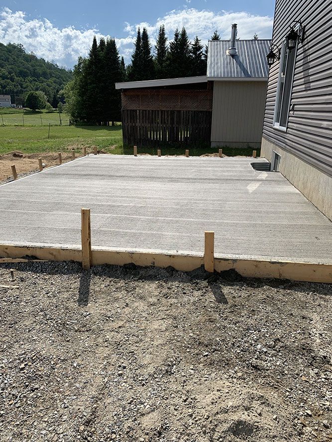 Construction d'une terrasse en béton à côté d'une maison à ossature bois et sol en gravier.