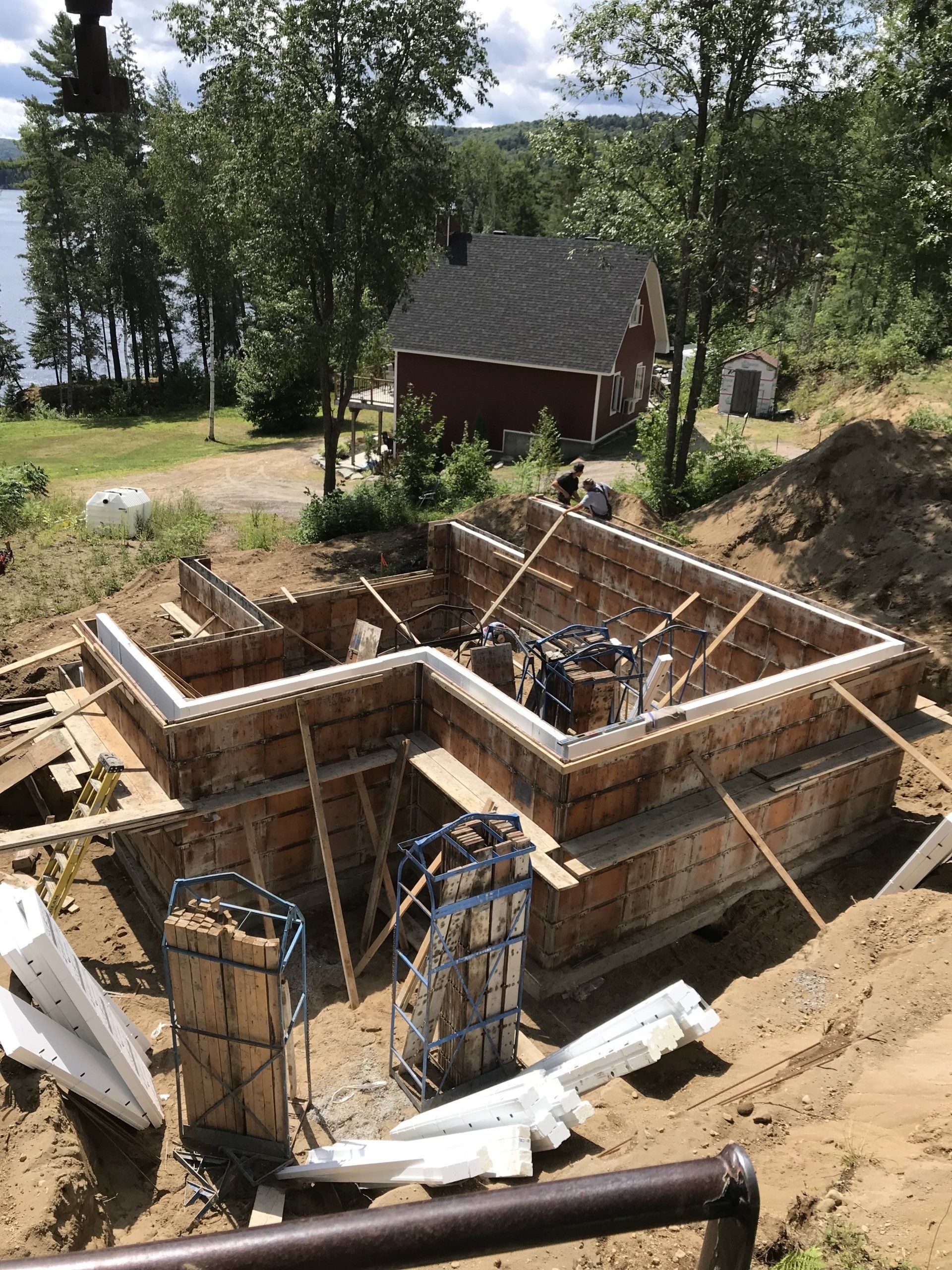Chantier de construction de fondations avec coffrages en bois, barres d'armature et un bâtiment rouge en arrière-plan.