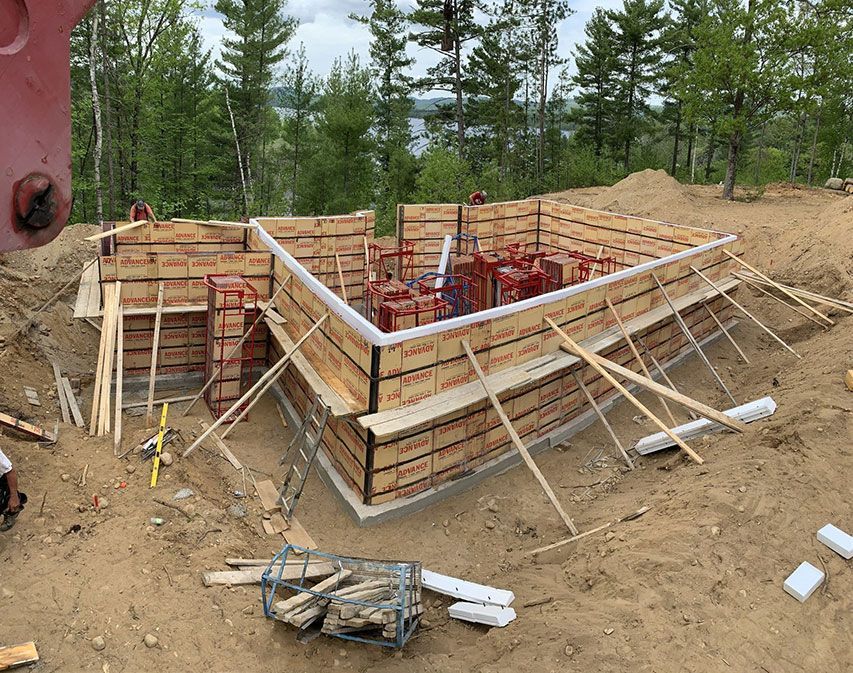 Coffrages de fondation en bois en cours de construction sur un terrain en terre ; supports, piquets et barres d'armature présents.