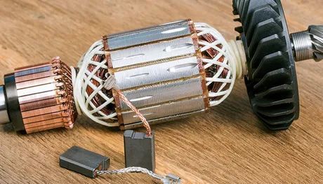 Disassembled power tool motor components: copper commutator, rotor, and carbon brushes on a wooden surface.