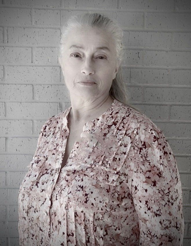 A person with light hair pulled back, wearing a floral button-up shirt, posing against a white brick wall.