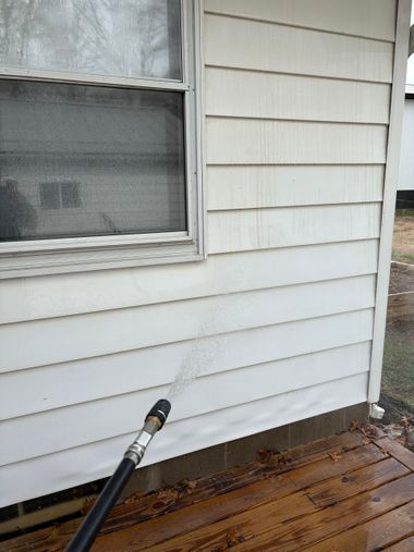A hand-held power washer sprays water onto a textured, beige stucco wall next to a black downspout.