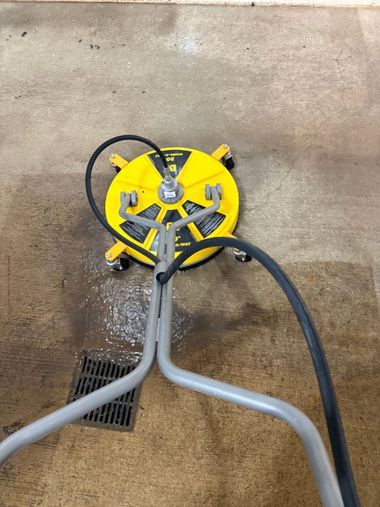 A person uses a pressure washer to clean a dark, stained concrete surface, revealing a light, clean area underneath.