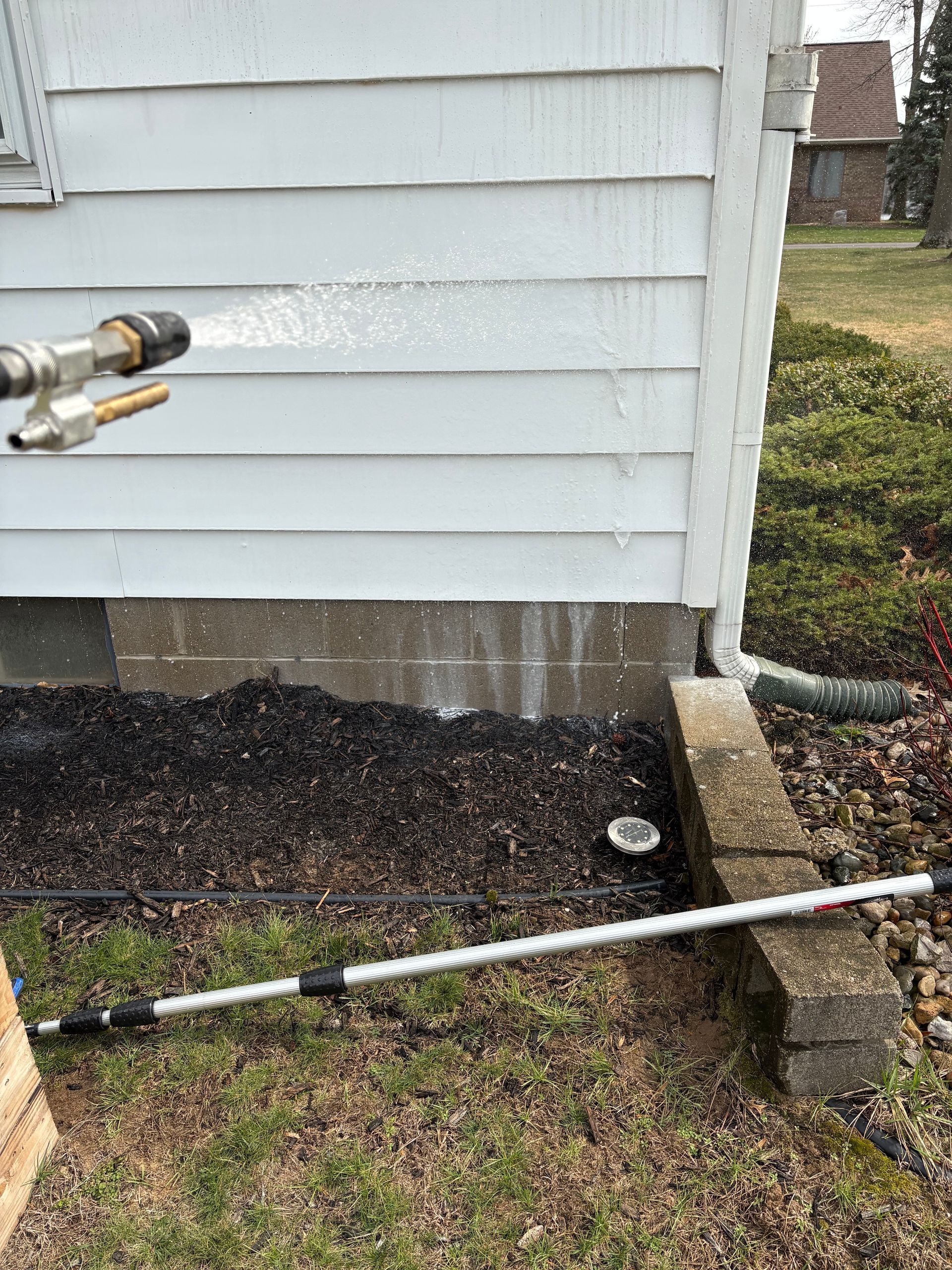 A person uses a pressure washer to spray cleaning foam onto the beige siding of a house above a dark gray brick wall.